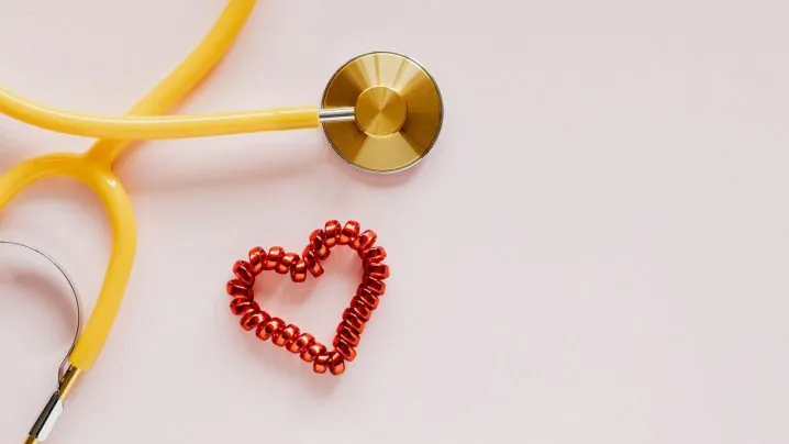 From above of yellow stethoscope near small red bead heart made for San Valentines Day on pale pink surface