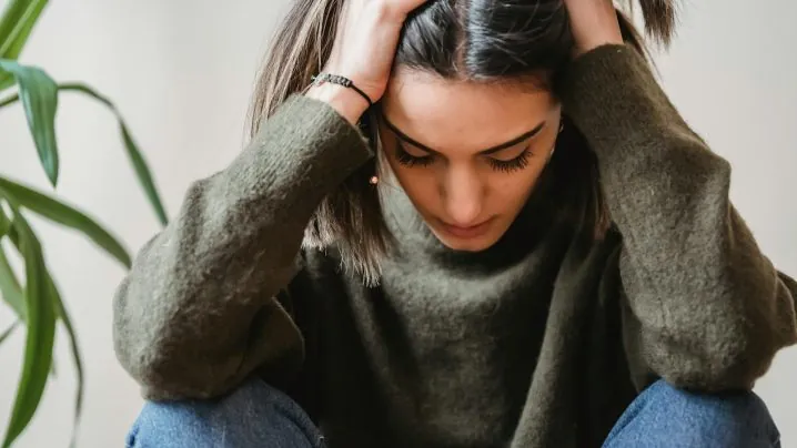 Crop disappointed young ethnic female with dark hair in casual clothes grabbing head with hands and looking down while sitting against white wall at home